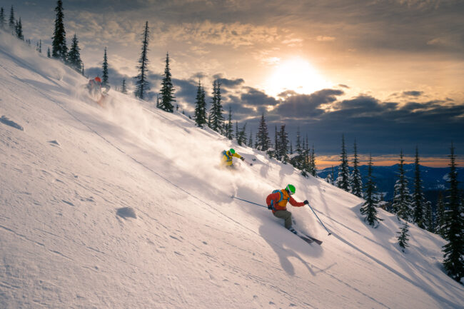 Two skiers making fresh tracks in freeride terrain as the sun rises over the clouds